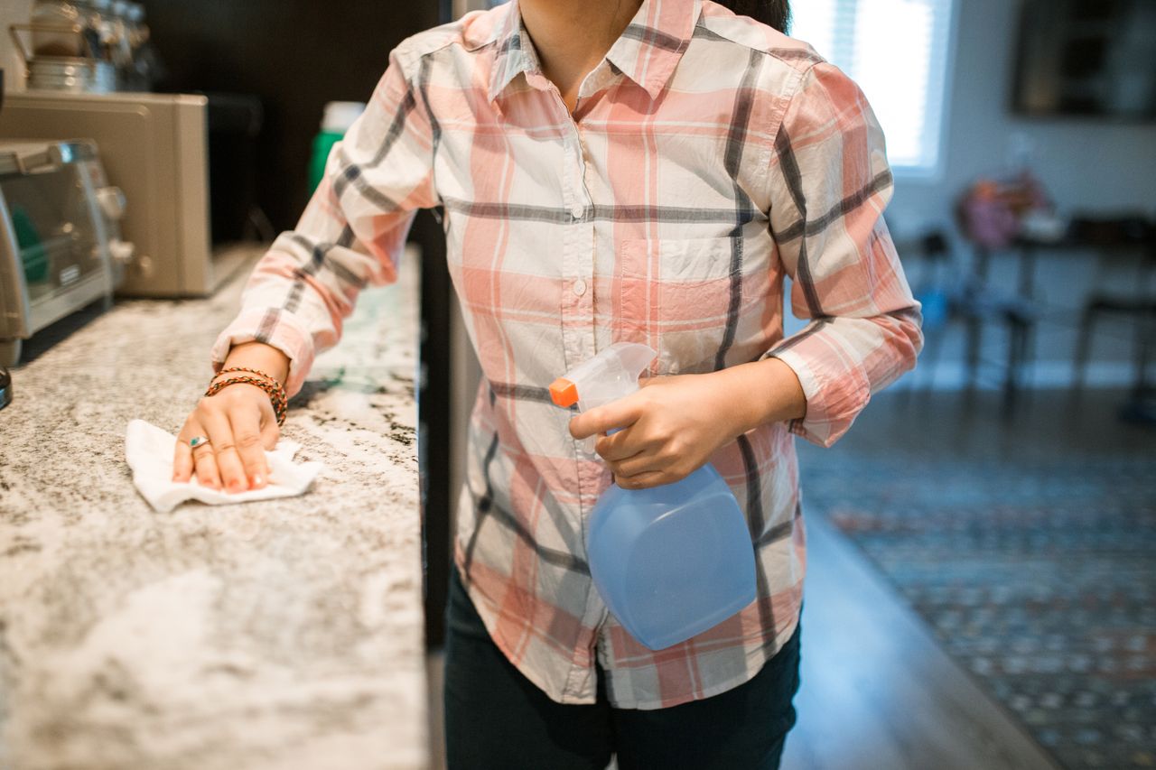 why-choose-us A woman wearing a plaid shirt cleans a kitchen counter using a spray bottle and cloth.
